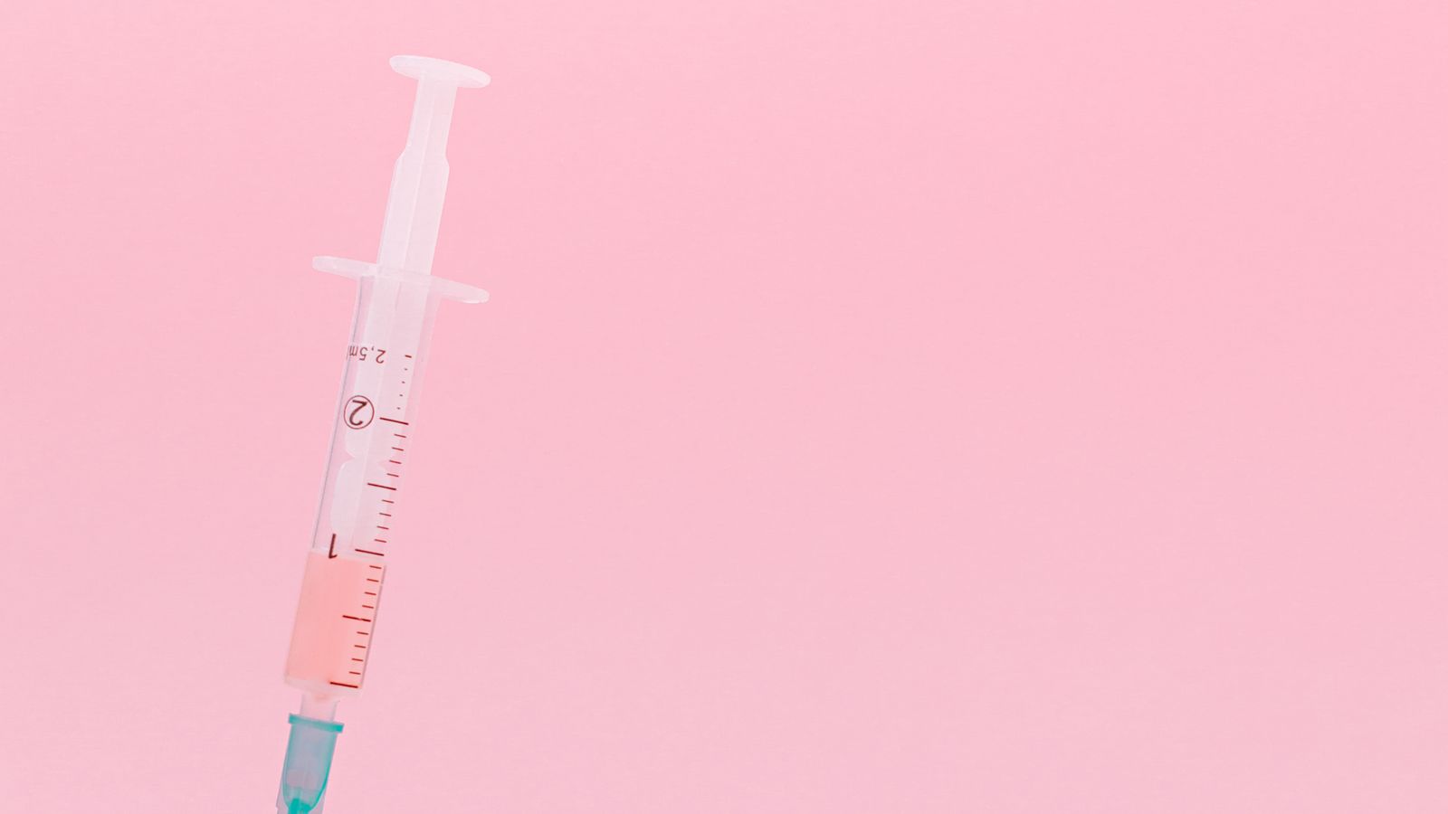A syringe draws liquid from a small vial resting on a pale pink surface.