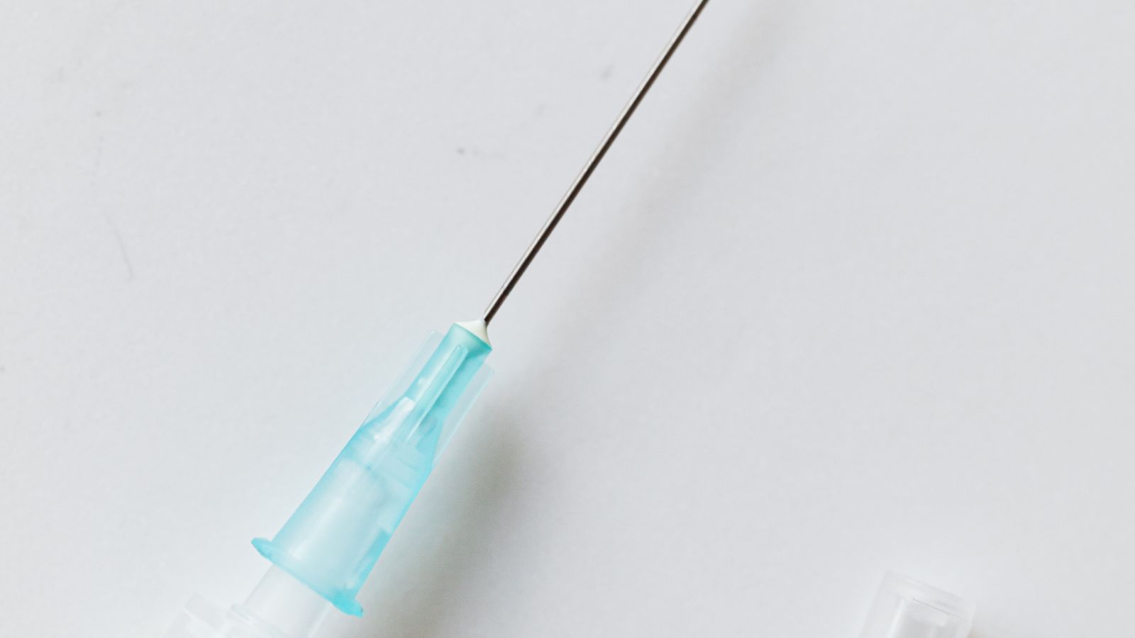 A capped medical needle rests on a white table beside a second syringe fitted with an exposed needle.