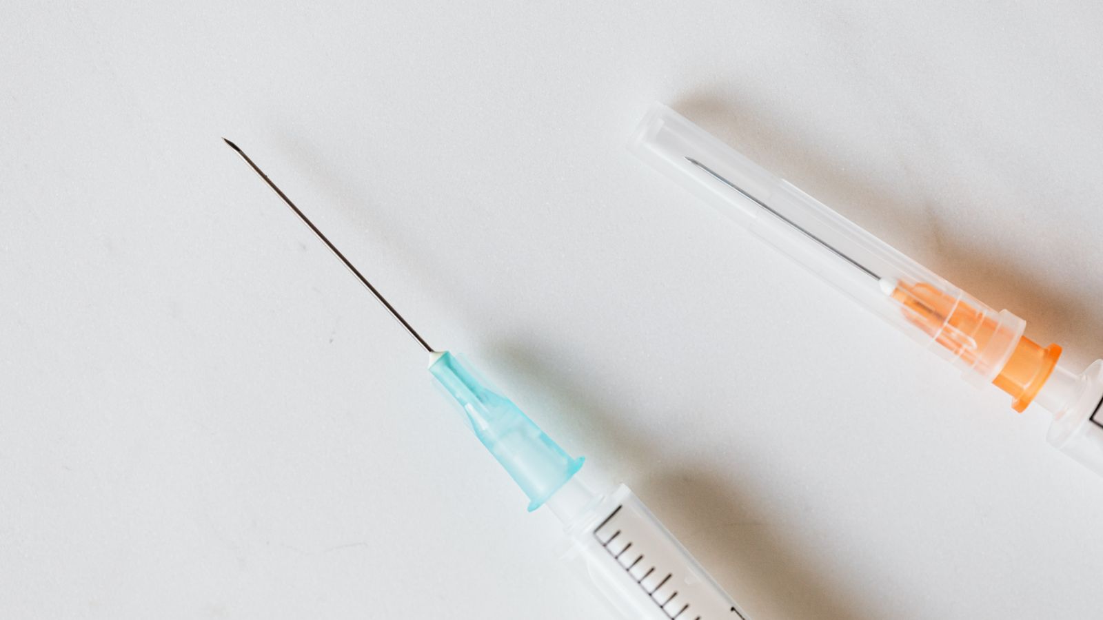 A medical syringe with its needle capped rests on a white table next to a second syringe fitted with an exposed needle.
